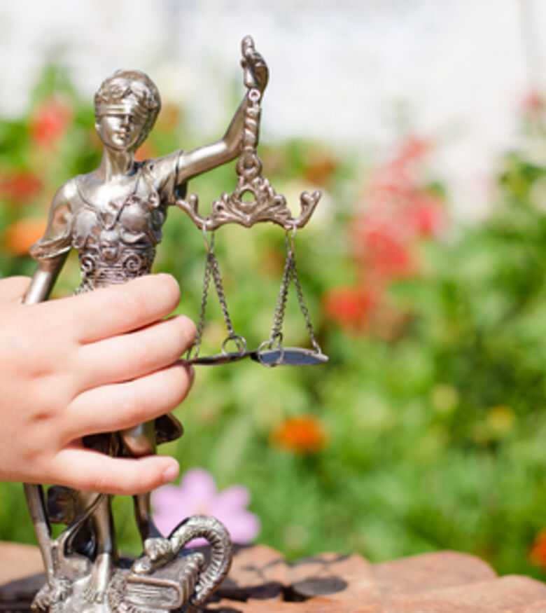 Hand holding a small statue of Lady Justice in front of a garden